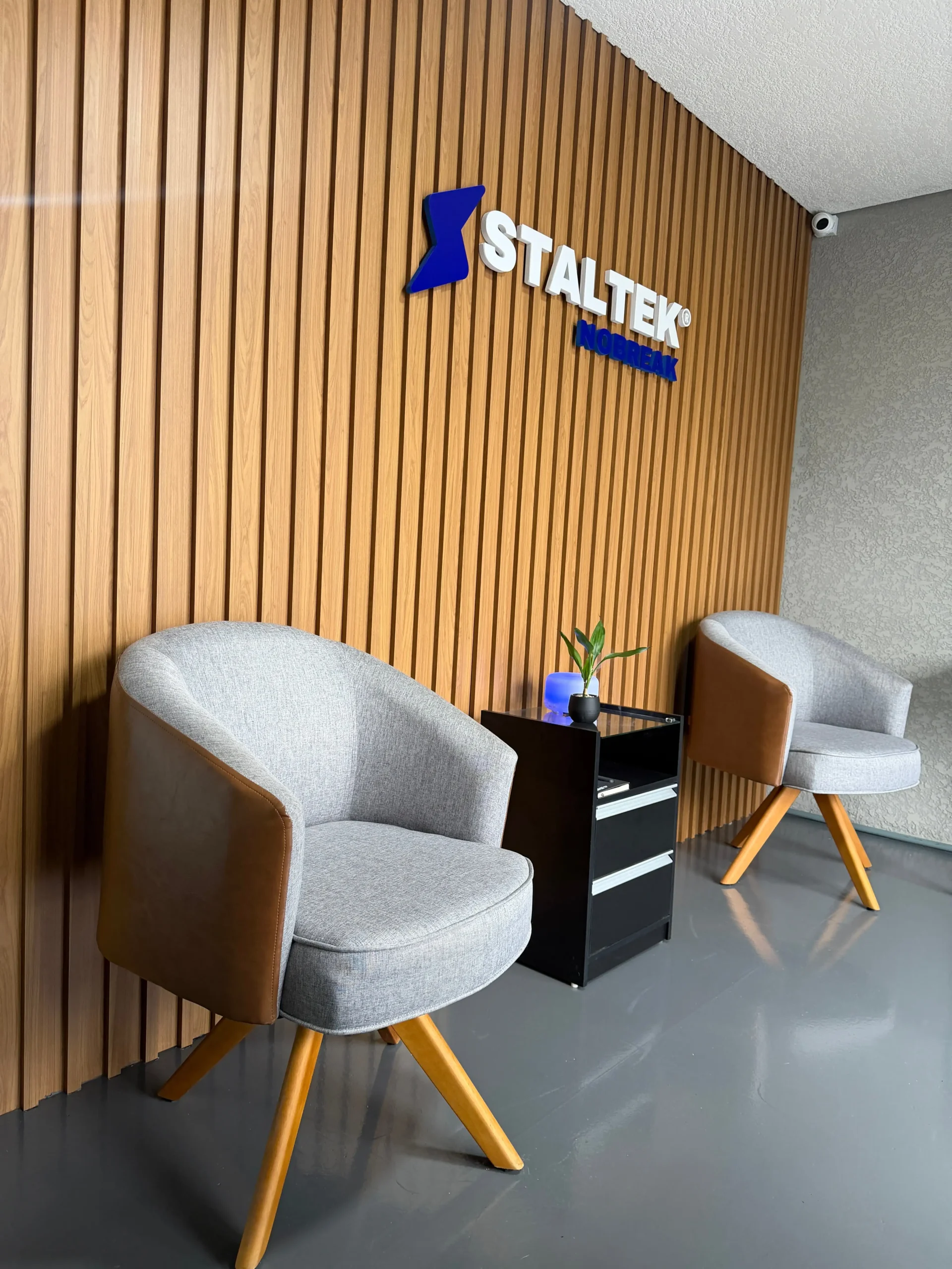 Área de recepção corporativa da Staltek Nobreak, apresentando um elegante painel ripado de madeira clara com o logotipo iluminado fixado, duas poltronas estofadas em cinza e marrom sobre um piso de resina cinza, e uma mesa lateral preta com um pequeno vaso e difusor de aroma.