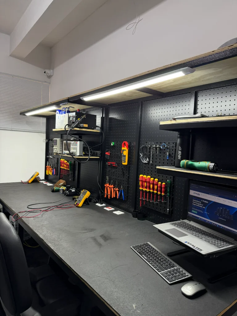 Bancada técnica da Staltek Nobreak organizada com ferramentas e instrumentos de medição Fluke, incluindo multímetro digital e alicate amperímetro, iluminada por LED, com um notebook exibindo o site institucional em primeiro plano.