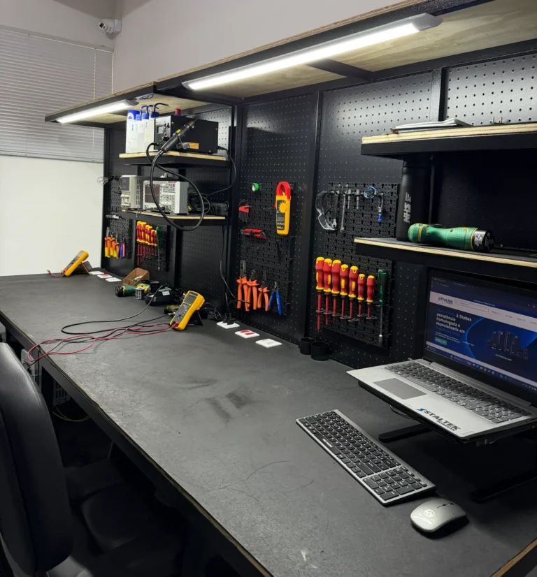 Bancada técnica da Staltek Nobreak organizada com ferramentas e instrumentos de medição Fluke, incluindo multímetro digital e alicate amperímetro, iluminada por LED, com um notebook exibindo o site institucional em primeiro plano.